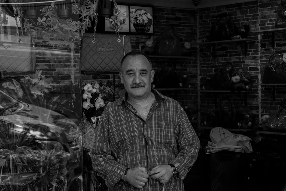 A man with a short beard and mustache, wearing a checkered shirt, stands inside a florist shop surrounded by an array of floral arrangements, hats, and decorative items. He is holding his hands togeth
