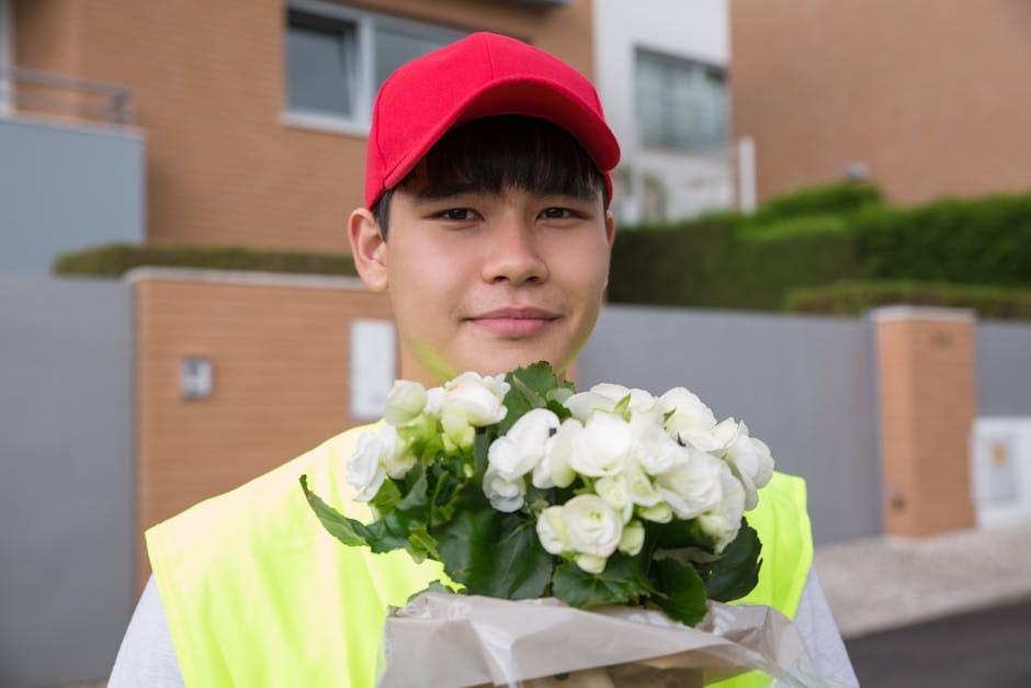 A young delivery person wearing a red cap and high-visibility yellow vest is standing outdoors in front of modern residential buildings, holding a bouquet of fresh white flowers, including roses and g