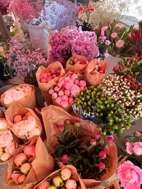 A vibrant display of freshly cut flowers and floral arrangement materials at a flower market. The scene features numerous wrapped bundles of pink peonies,牡丹, and other blooms, arranged in brown pa