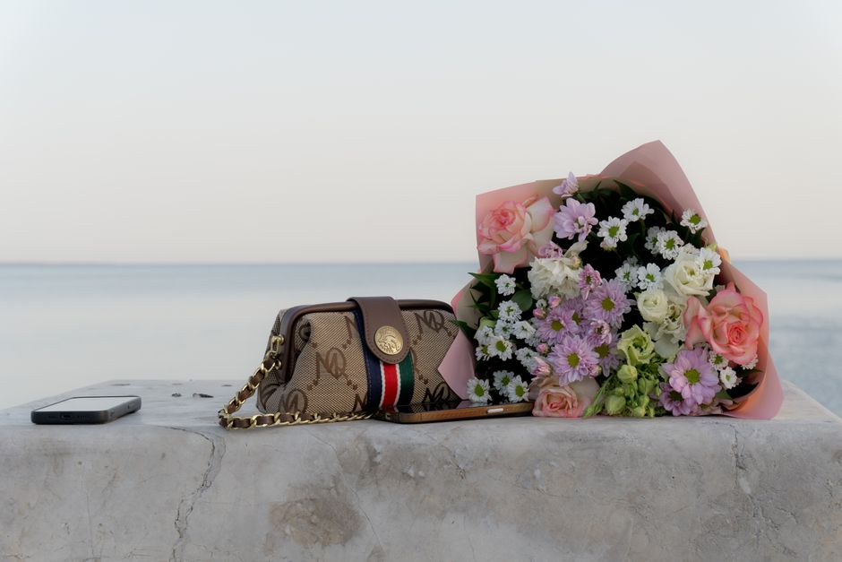 A bouquet of fresh flowers resting on a light grey stone surface, featuring pink roses, white daisies, and purple chrysanthemums wrapped in pink paper. Beside the bouquet is a designer handbag with a 