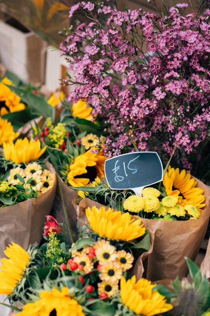 A vibrant display of freshly arranged flowers including bright yellow sunflowers, small white daisies with yellow centers, and various green foliage, all wrapped in rustic brown paper. Behind the bouq
