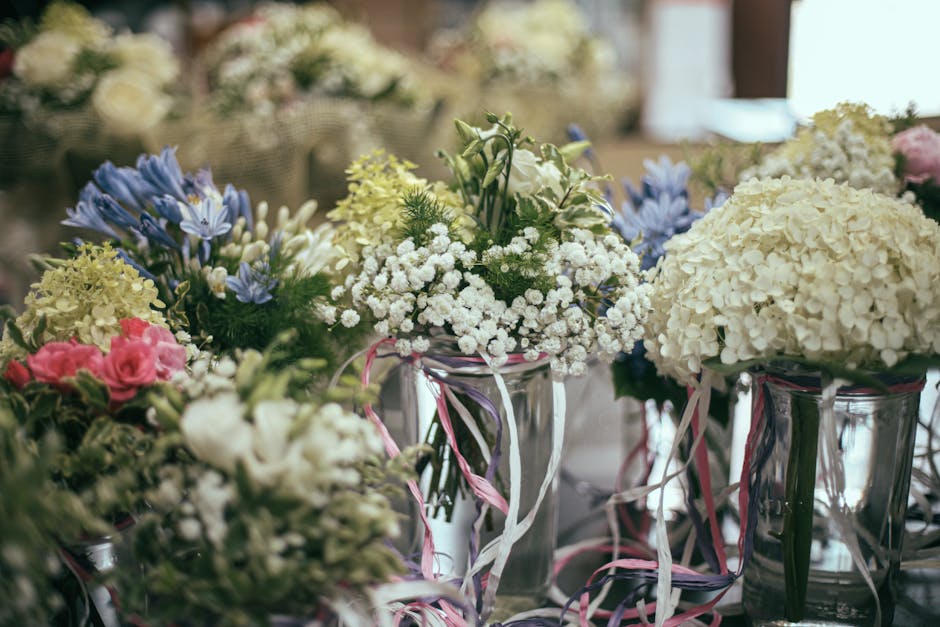 A close-up view of multiple floral arrangements in clear glass vases, featuring a variety of fresh flowers including white hydrangeas, pink roses, blue delphiniums, and white baby's breath, all with v