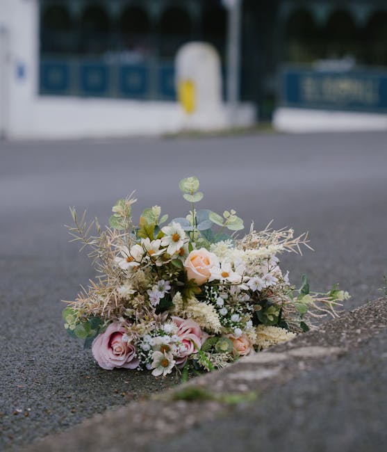 A delicate floral bouquet resting on a paved street, featuring soft pink roses, white daisies, cream-colored filler flowers, and sprigs of eucalyptus and other green foliage. The arrangement is loosel