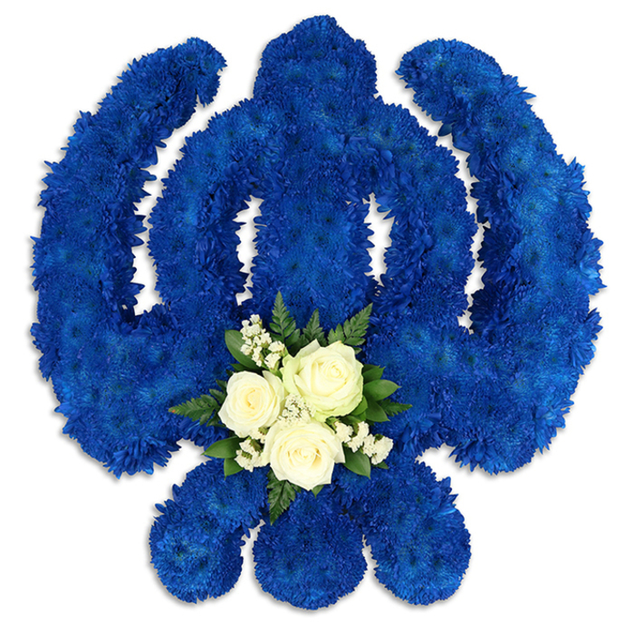 An impressive Sikh Khanda floral tribute dominates the image, carefully arranged so that every part of the sacred emblem is clearly defined against a crisp white backdrop. The central sword, the encircling chakkar, and the two symmetrical kirpans that curve gracefully on either side are all composed from compact royal blue chrysanthemums or similar petite blooms. These flowers are placed so close together that they form a dense, velvety surface, rich with texture and depth, creating a strong silhouette that feels both solemn and reassuring. The blue has a regal, uniform tone, reminiscent of deep twilight over the rooftops around Bounds Green, and it gives the piece a sense of spiritual gravity and calm. At the base of the central blade, a small yet prominent cluster of white and green flowers introduces a softer note. Three beautifully shaped white roses nestle at the centre, their petals gently unfolding in layered spirals that suggest tenderness and quiet devotion. They are surrounded by airy sprays of tiny white blossoms, likely gypsophila, which add a mist-like delicacy, and by lush dark green foliage that frames the cluster and offers a refreshing contrast to the intense blue. Gentle shadows along the edges of the Khanda hint at its thickness and structure, as though it has been built up on a solid base by an experienced florist who understands both the symbolism and the occasion. Ideal for Sikh funeral services and remembrance gatherings for families in Bounds Green and neighbouring areas, this design conveys profound respect, enduring faith, and a carefully considered tribute to a cherished life.