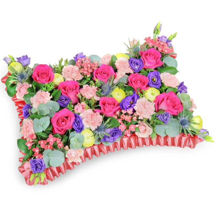 Captured at a modest oblique angle on a pure white backdrop, this rectangular floral pillow is a densely composed tapestry of colour and texture, consciously arranged to convey warmth and remembrance. The dominant notes come from numerous hot pink roses whose velvety petals catch the light and draw the eye across the cushion's surface. Intermingled pale blush carnations provide a soft, ruffled counterpoint, producing a sense of tactile abundance. Deep purple lisianthus punctuate the design with elegant, cup-like blooms, while lemon-yellow ranunculus or similar blossoms scatter bright, cheerful highlights that suggest celebration rather than sorrow. Silver-blue eryngium thistles are placed with compositional intent, their spiky silhouettes bringing a touch of wild, cool elegance and visual rhythm among the rounder blooms. Small clusters of pale pink filler blooms and varied green foliage - notably rounded, silvery eucalyptus leaves - fill pockets between larger flowers, creating depth and a cool aromatic undercurrent.

The perimeter is finished with a rich ruffled red satin ribbon that frames the cushion shape and lends the tribute a refined, classic character. Soft, even lighting reveals petal surface and shadow while preserving the arrangement's intimate, crafted feeling. This memorial pillow reads as an affirmation of life and cherished memories; I imagine it brought into local chapels or placed by family at a service in Bounds Green, Alexandra Palace events or Bowes Park, offering solace through colour, scent and considered floral artistry.