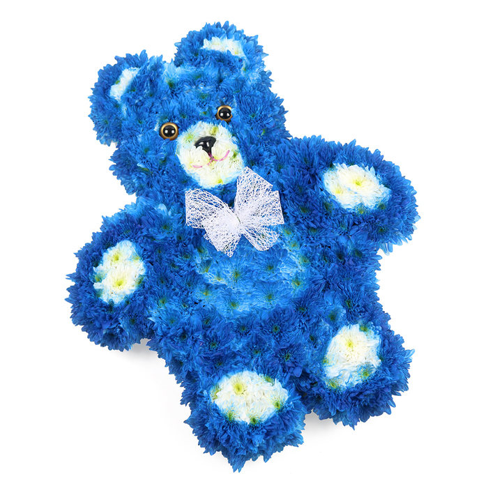 A tender floral tribute in the unmistakable shape of a teddy bear is shown lying flat on a plain white surface, every curve of its body meticulously built from fresh flowers. The bear's rounded head, tummy, arms, and legs are covered in a continuous layer of vibrant royal blue blooms, most likely small chrysanthemums or similar compact-headed flowers, arranged so closely that the texture resembles dense, velvety fur. These petals create gentle shading along the contours, making the bear appear softly sculpted rather than flat. Accents of creamy white and soft buttery yellow flowers form a circular muzzle, neat inner ears, and four oval pads on its paws, bringing a harmonious contrast that brightens the face and extremities. Two shiny, dark eyes and a small black triangular nose give the bear a serene, watchful expression, completed by a fine black line suggesting a subtle, reassuring smile. Around the neck, a sheer white bow-delicate and slightly opaque-rests with light ruffles, adding a sense of ceremony, as though the bear has been dressedespecially for a heartfelt goodbye. This arrangement feels perfectly at home in a quiet chapel near Bounds Green, or delivered to a family home off Green Lanes as a comforting focal point during a wake. The royal blue symbolises calm, remembrance, and sometimes the celebration of a baby boy's life, while the teddy shape evokes bedtime comfort and the safety of childhood. As an experienced florist in the area, I recognise the skill needed to keep the outline so clean and the expression so gentle, turning simple flowers into a deeply personal memorial.