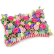 Viewed from a gentle, oblique angle against a clean white background, this pillow-shaped floral tribute reads like a soft, handcrafted memory. The rectangular 'cushion' is densely packed so each bloom nestles close to its neighbour: abundant hot pink roses with velvety, partially unfurling petals create the principal field of colour, while clusters of pale blush-pink carnations lend a ruffled, tactile surface that invites a comforting touch. Dashes of deep purple lisianthus punctuate the composition with bell-shaped depth, and sunny lemon-yellow ranunculus or buttercup-like blooms scatter bright, hopeful accents across the top. Architectural, silver-blue eryngium thistles introduce a cool, spiky contrast that balances the softness, and small sprays of delicate pink filler flowers add a whispered intricacy between the larger faces. Rounded, silvery-green eucalyptus leaves and other lush foliage weave through and beneath the flowers, their cool scent imagined even in the photograph and providing sculptural support and verdant depth.

The whole arrangement is edged in a rich, ruffled red satin ribbon that frames the cushion and defines its classic shape, giving the piece a formal, respectful finish. The lighting is soft and even, letting petal textures and subtle shadows show the careful symmetry and craftsmanship. Traditionally chosen as a sympathy pillow or memorial cushion, this design feels celebratory rather than sombre - a colourful, warm way to honour a life. As a local florist familiar with Bounds Green's quiet chapels and neighbourhood services, I picture this placed with care at a funeral or memorial in Bounds Green or nearby Bowes Park, offering solace and bright recollection where it is most needed.