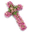 A serene and intricately crafted floral cross is set alone against a bright white background, its entire surface enveloped in a blend of soft pinks, mauves, and gentle purples that radiate quiet comfort. The cross appears full and three-dimensional, with flowers arranged so lavishly that they create a softly contoured, cushion-like effect. At the joining point of the horizontal and vertical arms, three fully open roses in muted dusty rose and light mauve form the emotional heart of the tribute, their layered petals unfolding like a calm breath. These roses are surrounded by lacy green fern fronds and deep green leaves, which add freshness and a slight woodland feel, balancing the pastel tones. Running along each arm and down the length of the cross are numerous lisianthus-style blossoms, their trumpet-shaped, subtly ruffled petals moving from bright to deeper pink and mauve, some with darker centers that create pockets of gentle shadow. Threaded among them are small clusters of delicate filler flowers in pink and purple, pale green buds, and tiny white blossoms that introduce lighter, airy moments within the more saturated tones. Around the outer edges, just visible beneath the dense blooms, lie slender ribbons or supporting elements in darker purple, lending definition and a hint of richness. The overall impression is one of peace, faith, and remembrance, the floral cross serving as a dignified Christian symbol for funeral tributes and memorials in Bounds Green. Whether placed by a coffin at a nearby chapel or brought to a graveside gathering, it offers a fragrant, beautifully crafted farewell, expressing love and respect when words are few.